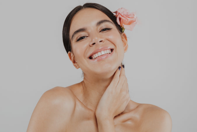 Woman with a flower in her hair smiling and touching her neck against a plain background