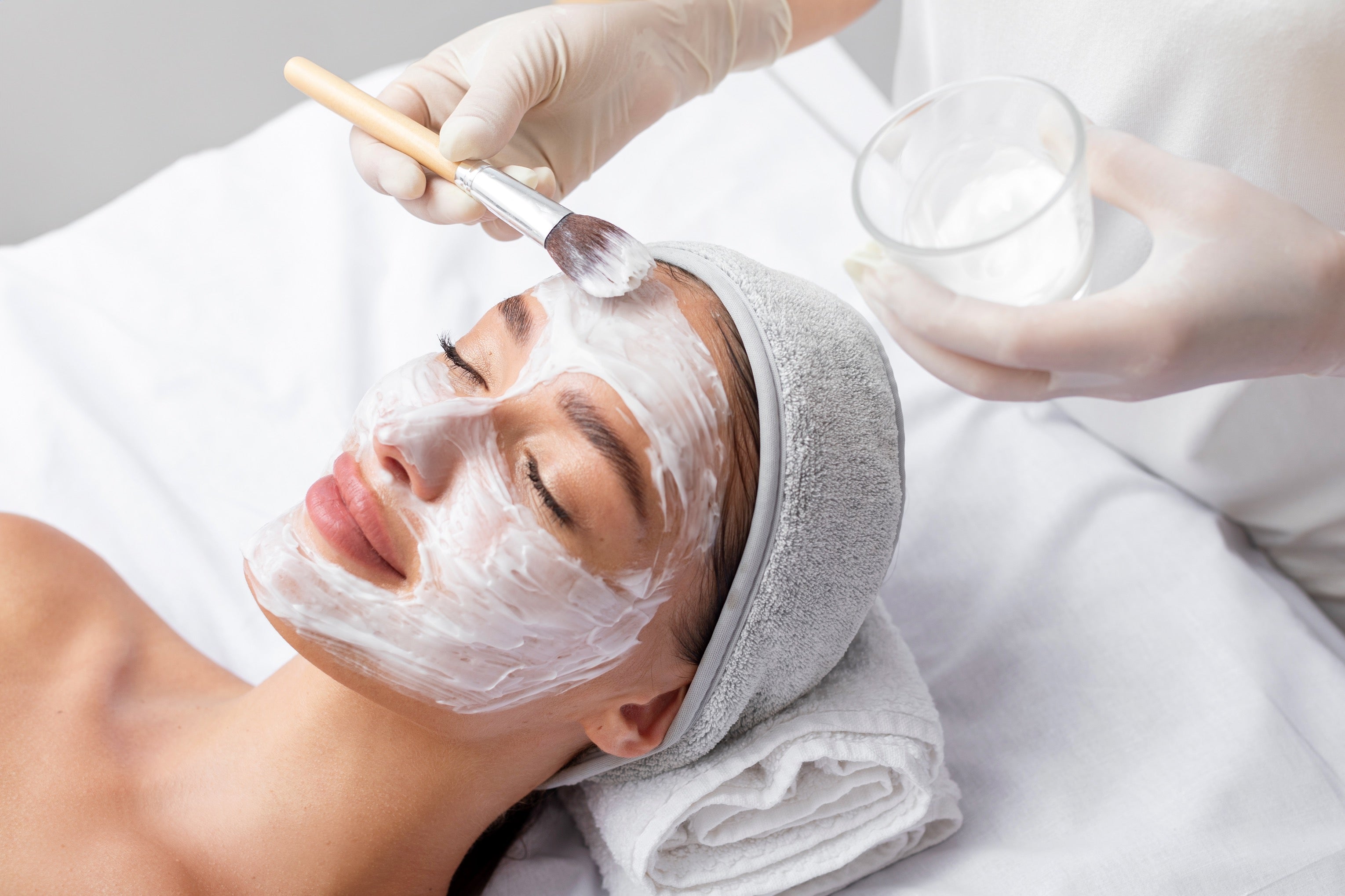 Person receiving a facial treatment with a brush and container of cream.